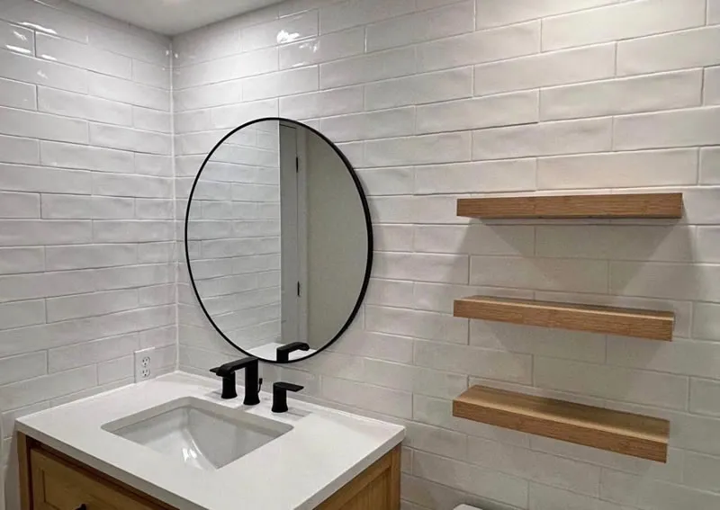 Vanity wall in glossy white tile with a round black-framed mirror and oak floating shelves